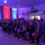 DSC_5686 Na zdjęciu grupa ludzi siedzi w sali z niebiesko-czerwonym oświetleniem. Wszyscy mają biało-czerwone wstążki i flagi. Tło jest jasne, z ozdobnymi zasłonami.