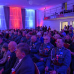 DSC_5689 Sala pełna ludzi w mundurach i formalnych strojach. Niebiesko-czerwone światła oświetlają scenę. Uczestnicy trzymają polskie flagi.