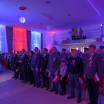 DSC_5690 Grupa osób w eleganckich strojach stoi w dużej sali. Dominuje oświetlenie w kolorach niebieskim i czerwonym. Uczestnicy mają przypięte biało-czerwone kokardy.