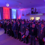 DSC_5692 Grupa osób stoi na uroczystości, ubrana w patriotyczne stroje. Tło podświetlone jest na czerwono, biało i niebiesko. Osoby trzymają małe flagi Polski.