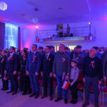 DSC_5695 Grupa ludzi stoi w sali oświetlonej na czerwono-niebiesko. Uczestnicy mają biało-czerwone ozdoby i trzymają polskie flagi. W tle balkon z obserwatorami.