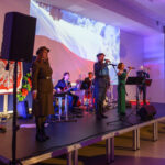 DSC_5696 Na zdjęciu jest grupa muzyków na scenie, otoczeni flagą Polski. Tło wypełnia biało-czerwona flaga i projekcja. Muzycy noszą eleganckie stroje, scena jest oświetlona na fioletowo.