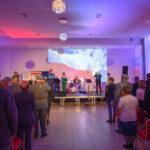 DSC_5698 Na zdjęciu jest koncert z widownią, na scenie zespół. Światła w kolorach czerwono-białych, w tle projektor z flagą. Sala jest przestronna z krzesłami.