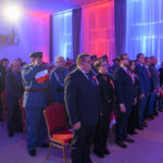 DSC_5699 Grupa ludzi stoi w eleganckiej sali, ubrani w formalne stroje, z biało-czerwonymi flagami. Tło oświetlone jest na niebiesko i czerwono. Wydarzenie ma oficjalny charakter.