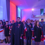 DSC_5703 Grupa ludzi ubrana odświętnie z polskimi flagami i kotylionami. Tło oświetlone w kolorach biało-czerwonych i niebieskich. Sala z wysokim sufitem i balkonem.