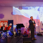 DSC_5704 Na scenie mężczyzna przemawia, w tle wyświetlona flaga Polski. Obok ustawione są instrumenty muzyczne i sprzęt nagłaśniający. Oświetlenie w ciepłych kolorach.