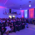 DSC_5715 Sala pełna ludzi siedzących na uroczystości, z kolorowym oświetleniem w niebieskich i czerwonych tonach. W tle widoczne są zasłony i scena z oświetleniem. Uczestnicy trzymają polskie flagi.