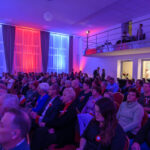 DSC_5717 Grupa ludzi siedzi w sali oświetlonej na czerwono, niebiesko i biało. Tło stanowią zasłony w ciemnych odcieniach. Na piętrze są widoczni technicy przy sprzęcie.