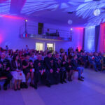 DSC_5725 Na zdjęciu widać dużą salę wypełnioną ludźmi. Ściany są oświetlone na niebiesko i czerwono. Uczestnicy siedzą na krzesłach, niektórzy trzymają polskie flagi.