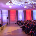 DSC_5756 W sali koncertowej osoby siedzą na krzesłach z biało-czerwonymi flagami. Scena oświetlona jest fioletowo i czerwono. Na scenie stoją wykonawcy w eleganckich strojach.