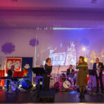 DSC_5759 Na scenie zespół muzyczny, tło z polskim godłem i zdjęciem kościoła. Kolorowe oświetlenie sceniczne w tonach fioletu i czerwieni. Uczestnicy w eleganckich strojach.