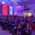 DSC_5767 Na zdjęciu widzimy salę z tłumem ludzi trzymających małe flagi Polski. Oświetlenie w kolorach: niebieskim, czerwonym i białym. Tło to sceniczne dekoracje i balustrada na drugim planie.