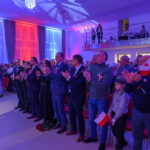 DSC_5774 Grupa ludzi stoi i bije brawo; sala w biało-czerwonych światłach. Wiele osób trzyma polskie flagi. W tle balkon z obserwatorami.