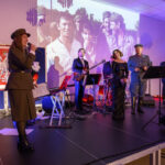 DSC_5775 Na scenie zespoły w eleganckich strojach wojskowych oraz cywilnych, z instrumentami. Tło zawiera biało-czerwoną flagę i projekcję archiwalnych zdjęć. Oświetlenie w ciepłych odcieniach fioletu.