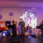 DSC_5784 Na scenie zespół muzyczny z instrumentami, wokalistą w mundurze. Tło: biało-czerwona flaga, czarno-białe zdjęcie pary. Kolorowe światła tworzą atmosferę koncertu.