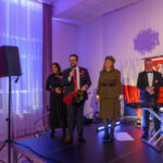 DSC_5789 Grupa ludzi stoi na scenie, jeden z nich trzyma mikrofon. W tle są biało-czerwone dekoracje. Scena jest oświetlona fioletowym światłem.