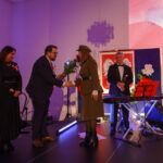 DSC_5797 Na scenie mężczyzna wręcza kobiecie kwiaty; w tle flaga z orłem. Dominuje fioletowe oświetlenie. Obok stoją muzycy z instrumentami.