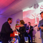 DSC_5801 Na scenie widać ludzi w eleganckich strojach, trzymających czerwone róże. Tło jest jasne z napisem „Święto Niepodległości 11 listopada”. Panuje uroczysta atmosfera z kolorowym oświetleniem.