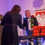 DSC_5805 Mężczyzna w smokingu trzyma różę i podaje rękę kobiecie na tle godła Polski. Kolory dominujące: czerń i czerwony. W tle jasne ściany.