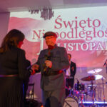 DSC_5812 Na zdjęciu widać wydarzenie z okazji Święta Niepodległości. Osoba w mundurze wręcza różę, w tle bęben i napis. Kolory dominujące to czerwony i biel.