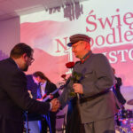 DSC_5816 Mężczyźni w garniturach i mundurze podają sobie dłonie na tle sceny. Dominują kolory czerwony, szary, niebieski. W tle napis Święto Niepodległości.