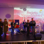 DSC_5831 Grupa osób na scenie celebruje Święto Niepodległości 11 listopada. Tło to biało-czerwony plakat z orłem. Scena oświetlona fioletowym światłem.