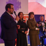 DSC_5833 Grupa ludzi na scenie, kobieta w mundurze trzyma różę. Tło z flagą Polski, ciepłe oświetlenie. Mężczyzna z mikrofonem.