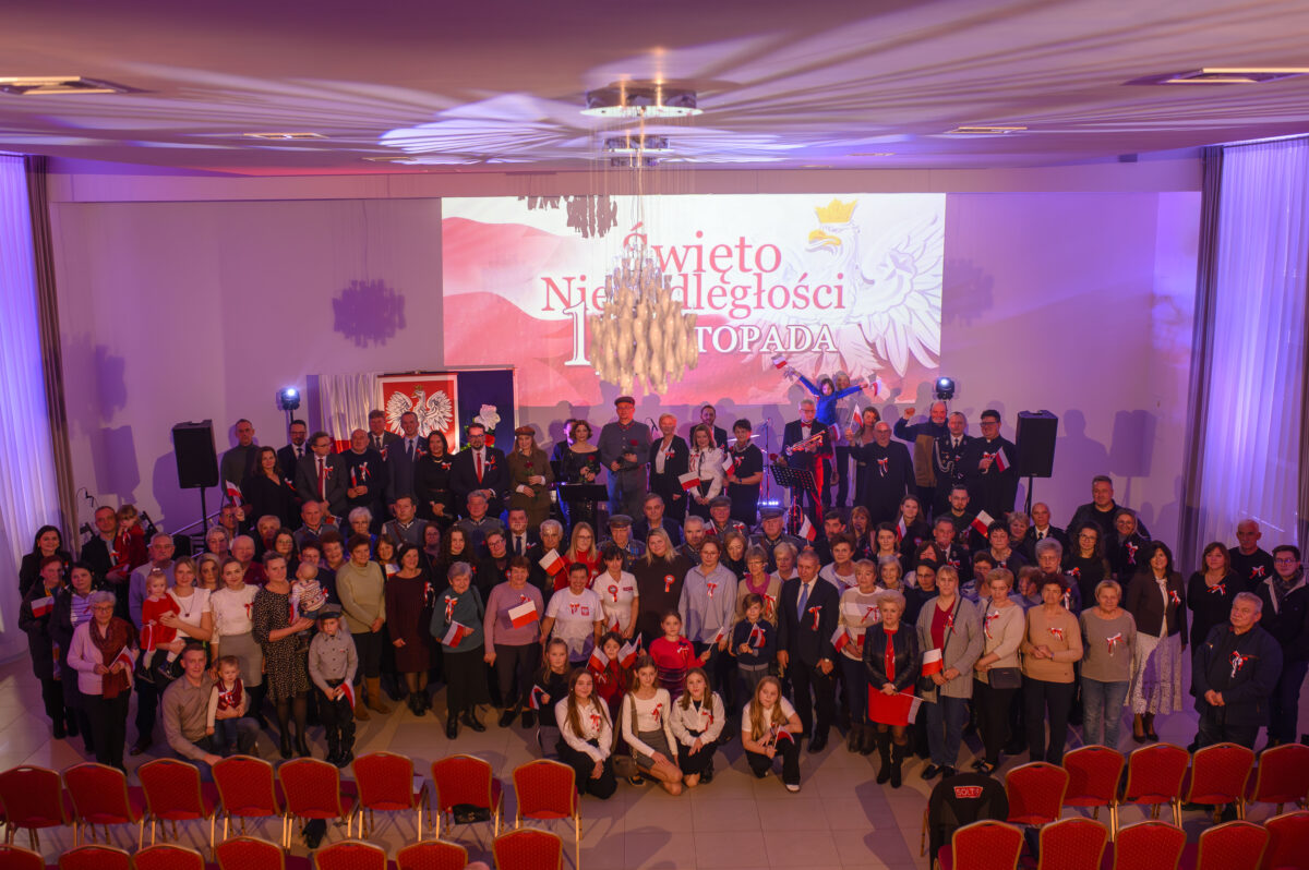 Grupa ludzi stoi na tle biało-czerwonej sceny z napisem Święto Niepodległości. Ubrani w odświętne stroje, trzymają flagi Polski. W tle widać duży żyrandol i ekran z orłem.