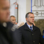 DSC_7728 Na zdjęciu widoczny jest mężczyzna w ciemnym płaszczu. W tle inne osoby oraz rozmyte dekoracje na ścianie. Zdjęcie jest kolorowe.