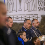 DSC_7730 Na zdjęciu jest grupa ludzi w mundurach i dzieci, w tle ściana z rysunkami. Przeważają kolory niebieski i szary. Scena ma oficjalny charakter.