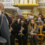 DSC_7734 Muzycy orkiestry dętej grają w kościele. Ubrani w granatowe mundury z czerwonymi akcentami na tle ozdobnych ścian. Klarneciści siedzą z przodu, inni z tyłu mają instrumenty dęte.