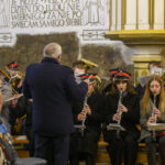 DSC_7736 Orkiestra dęta w kościele, muzycy w ciemnych uniformach i czerwonych czapkach. Dyrygent w ciemnym płaszczu na środku. Ściany jasne, zdobione napisami i ornamentami.