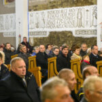 DSC_7739 Na zdjęciu jest grupa ludzi w kościele. Wnętrze jest jasne, z drewnianymi ławkami i białymi ścianami z napisami. Osoby siedzą, niektóre trzymają flagi.