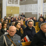 DSC_7741 Ludzie siedzą w kościele, skupieni na wydarzeniu. Ubrani są w ciemne płaszcze, niektórzy trzymają polską flagę. W tle jasne ściany z dekoracjami.