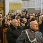 DSC_7743 Na zdjęciu widać tłum ludzi siedzących w drewnianych ławkach w kościele. Dominują kolory ziemi i szarości, część osób ma na sobie ciepłe ubrania. Tło zdobią białe ściany z dekoracyjnymi napisami.