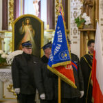 DSC_7755 Grupa osób w mundurach stoi przed obrazem Jezusa Miłosiernego w kościele. Widoczne są flagi w kolorach czerwonym i niebieskim oraz białe kwiaty. Tło zdobią złote elementy i napisy na ścianie.