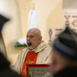 DSC_7763 Mężczyzna w kościelnych szatach mówi do mikrofonu. Jasne tło z figurą świętą. Kolory szaty: złoty i czerwony.