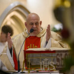 DSC_7772 Na zdjęciu ksiądz w złotej szacie czyta przy ołtarzu. W tle widoczne są łuki i ściana w ciepłych barwach. Obok otwarta księga i mikrofon.