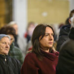 DSC_7776 Na zdjęciu są skupieni ludzie w pomieszczeniu, przeważają stonowane kolory. Kobieta na pierwszym planie nosi bordowy płaszcz. W tle są inne osoby, ubrania w zimowej kolorystyce.