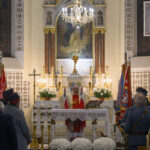 DSC_7783 Ksiądz prowadzi mszę w kościele z jasnym ołtarzem i dużym złotym żyrandolem. Białe kwiaty zdobią ołtarz. W tle widoczne są barwne flagi i wierni.