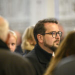 DSC_7793 Zdjęcie przedstawia mężczyznę w okularach wśród tłumu. Dominują kolory stonowane, głównie szarości. Tło jest rozmyte, co skupia uwagę na postaci.