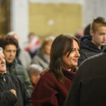 DSC_7798 Na zdjęciu widać grupę ludzi w pomieszczeniu. Kobieta na pierwszym planie jest ubrana w bordowy płaszcz, a inni mają zimowe ubrania. Tło to rozmyta ściana w jasnych kolorach.