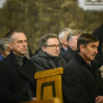 DSC_7803 Grupa ludzi siedzi w drewnianych ławkach w kościele. Tło jest kamienne, w stonowanych barwach. Osoby wyglądają na zamyślone.