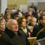 DSC_7804 Grupa ludzi w kościelnych ławach, patrzących w tym samym kierunku. Dominują kolory beżowe i brązowe. Tło jest rozmyte, skupia uwagę na postaciach.