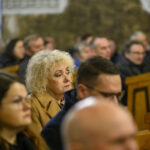 DSC_7806 Grupa ludzi siedzi w kościele, skupiona na nieznanym wydarzeniu. Kobieta w jasnym płaszczu na pierwszym planie. Tło rozmyte, ciepłe odcienie.