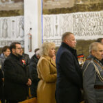 DSC_7808 Grupa osób w kościele ubrana formalnie stoi, w tle tablica z napisem i aniołami. Dominują kolory ciemne. Po prawej stronie widać mężczyznę w mundurze.