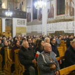 DSC_7814 Grupa ludzi w jasnym kościele siedzi na drewnianych ławkach. Ściany ozdobione malowidłami, z białymi kolumnami. Światło żyrandola rozprasza się po całym pomieszczeniu.