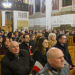 DSC_7816 Grupa ludzi w kościele siedzi na drewnianych ławkach. Tło zdobią jasne malowidła i kolumny. Atmosfera jest poważna i skupiona.