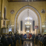 DSC_7821 Wnętrze kościoła z licznymi wiernymi i sztandarami. Dominuje złoto, beż i czerń; sufit drewniany, tło to ołtarz. Centralnie wiszący żyrandol.