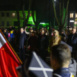 DSC_7831 Grupa osób stoi na zewnątrz w nocy, trzymając polskie flagi. Tło oświetlone zielonym światłem, wokół drzewa. Przód zdjęcia zajmuje flaga Polski.